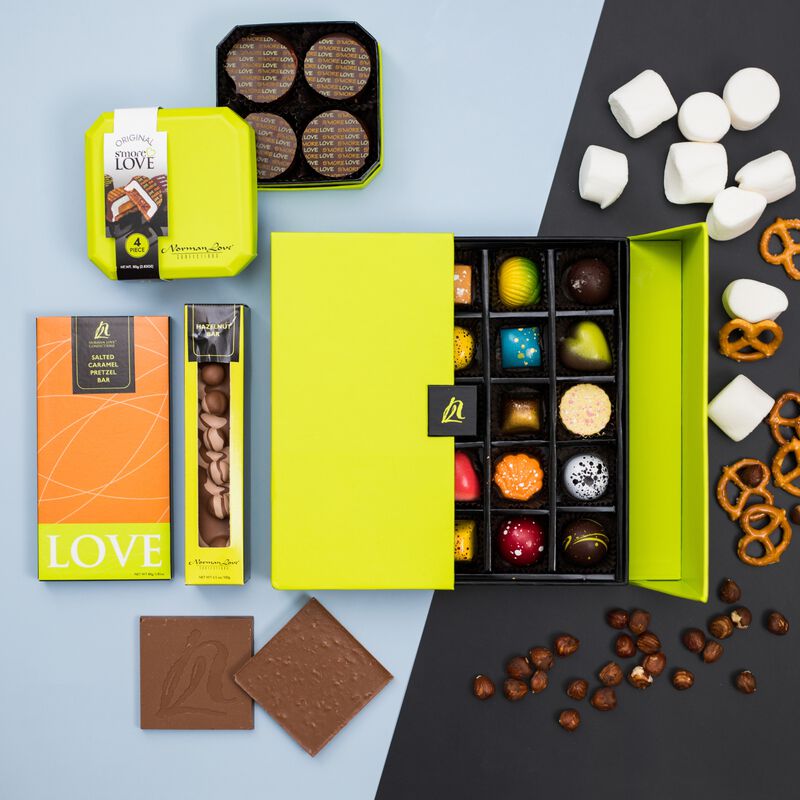 Overhead view of half open lime green signature box displayed with Box of S'mores Love, Hazelnut Chocolate Bar and Salted Caramel Pretzel Inclusion Bar to the left. Marshmallows, pretzels and hazelnuts curve around truffle box.